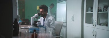 Gözlüklü ve koruyucu eldivenli genç bir bilim adamı mikroskop altında laboratuvarda bir tüp tutuyor. Tıp, biyoteknoloji, kimya.