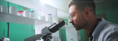 Mikroskop altında gözlüklü ciddi bir bilim adamı ışık laboratuarında bir tüp tutuyor. Tıp, biyoteknoloji, kimya.