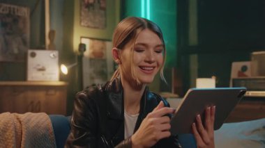 Siyah deri ceketli, şirin, şık, beyaz bir kadının kanepede oturup tablette video izlediği bir fotoğraf. Çekici, kaygısız bayan hippi akşamları kapalı alandaki cihazlara dokunuyor.