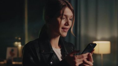 Siyah deri ceketli, akıllı telefondan mesaj atan mutlu, beyaz bir kadının portresi. Uzun sarı saçlı çekici bir kız. Kapalı alanda flört uygulamasında sohbet ediyor. Gece vakti. Arka planda neon ışıkları