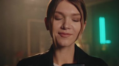 Çıplak makyajlı neşeli, genç ve çekici bir kadın akıllı telefon ekranına bakıyor ve mesaj okuyor. Kapalı mekanda şık giyinmiş güzel bir kızın yakın plan portresi. Oturma odasının bulanık arka planı