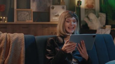 Çekici, mutlu, genç bir kadın koltukta oturup gülümseyen ve evinde dinlenen tablet bilgisayar kullanıyor. Gece vakti.