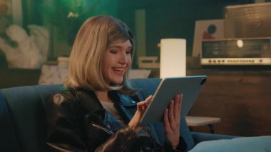 Çekici genç kadın tablet bilgisayar kullanıyor koltukta oturuyor gülümsüyor ve evde rahatlıyor. Gece vakti.