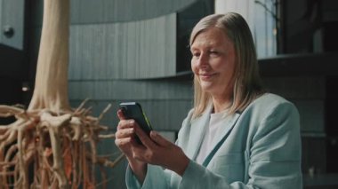 Sarışın, başarılı iş kadınının akıllı telefondan mesajlaşması. Zarif kıyafetli güzel, mutlu bir kadın modern otel salonuna bakıyor. Gündüz vakti. Kapalı, 60 'lı yaşlarda