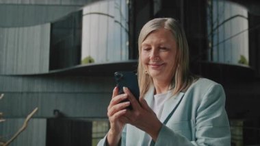 Düşünceli, kendine güvenen, 60 'lı yaşlarda gülümseyen, akıllı telefon kullanan ve kocasıyla sohbet eden bir kadının görüntüsü. Güzel tasarlanmış otel odasında duran zarif mavi ceketli ve büyük küpeli çekici moda işkadını.