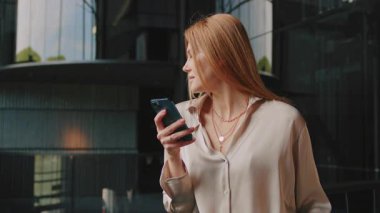 Modern otel salonunda dururken zarif bluz giymiş kızıl saçlı, gülümseyen bir iş kadınının akıllı telefonuyla sohbet etmesi. İç mekanda kendine güvenen güzel bir kadın.