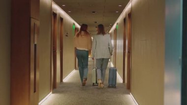 Modern otel koridorunda yürüyen zarif kıyafetler içindeki zayıf, kızıl saçlı ve sarışın iki iş kadınının arka planı. Kadınlar bavullarını yuvarlıyor. Harika bir iç mekan. Mesafe atışı. Zarafet ve lüksün atmosferi
