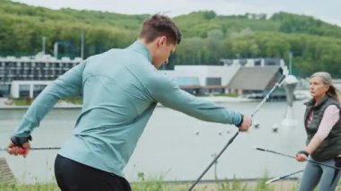 Genç antrenör, İskandinav yürüyüş çubuklarını göllü yeşil orman parkında nasıl kullanacağını gösteriyor. Bulutlu hava