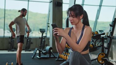 Spor giyimli düşünceli çekici bir kadın antrenman arasında bankta oturuyor ve akıllı telefondan sohbet ediyor. Formda, güzel esmer, geçmişi bulanık at kuyruklu. Sağlık, spor konsepti. Kapalı