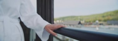 Kapalı çekimde, büyük küpeli, güzel gri saçlı olgun bir kadının spa merkezindeki otel odasının balkonundan bakışları görülüyor. Harika bir manzara. Anamorfik mercekle çekilmiş. Sağlıklı bir yaşam tarzı. Gündüz