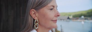 Kameraya bakan beyaz saçlı çekici olgun bir kadının yakın plan portresi. Büyük küpeli gülümseyen bayan otel odasının balkonundan panoramik manzaranın keyfini çıkarıyor. Anamorfik lens