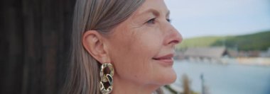 Beyaz saçlı, güzel küpe takan ve otel balkonundaki kameraya bakan güzel görünümlü beyaz kadın portresi. Arka plandaki spa merkezinin bulanık görüntüsü. Yakın çekim.