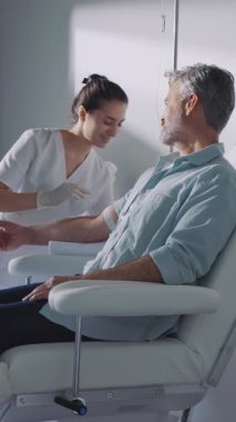 Kapatın genç bayan hemşire hazırlanıyor. Oturan hasta adamdan kan alın. Konuşurken ışık odasında ve modern klinikte gülümseyin. Hastanedeyiz. Yavaş çekim