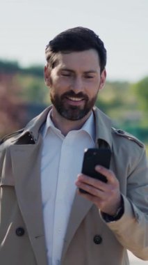 Palto giyen mutlu, şık, yakışıklı iş adamı göl kenarındaki parkta akıllı telefon kullanıyor. Gülümseyerek etrafına bak. Kapatın. Yavaş çekim