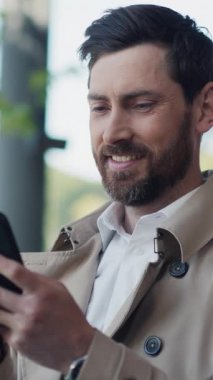 Paltoyla sakallı, genç, şık, yakışıklı bir adam güneş ışığında akıllı telefon standı kullanıyor. Kapatın. Yavaş çekim