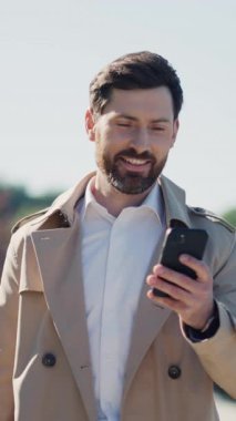 Ciddi, şık, ceketli yakışıklı iş adamı portresi güneş ışığında gölün yanındaki parkta akıllı telefon standı kullanıyor. Kapatın. Yavaş çekim