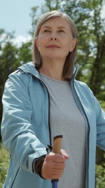 Sopalarıyla yürüyen muhteşem, sportif yaşlı bir kadının görüntüsü. Spor elbiseli çekici bir kadının portresi doğada İskandinav yürüyüşü yapıyor. Otel kompleksi. Sağlıklı eğitim, yaşam tarzı. Rahatlatıcı, huzurlu, dinlendirici, açık havada