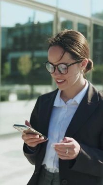 Zarif, kendine güvenen, zarif, resmi takım elbiseli beyaz bir kadının akıllı telefonu elinde tutarak etrafa bakındığı görüntüler. Bun saç stiliyle gülümseyen zeki iş kadınının portresi. Arka plan bulanık. Açık havada.