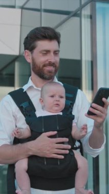 Yaklaş genç yakışıklı baba kucağında kucağında küçük bir bebekle geziyor ve sokakta gülümseyen telefon daktilosu kullanıyor. Dolayısıyla sırt çantası. Ebeveynler. Gadget. Akıllı telefonmuş. Cep telefonu. Yavaş çekim