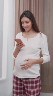 Güzel genç esmer kadın pencerenin yanında akıllı telefon kullanıyor. Hamile genç bir anne çocuk için internetten bebek alışverişi bekliyor..