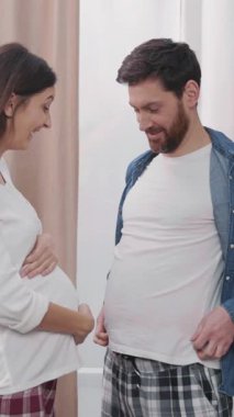 Güzel, genç bir kadın hamile kocasına gülen bir çocuk bekliyor. Komik adamın karnını göstermesi. Aileler eğlencelidir.