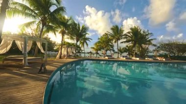 Palmiye ağaçlarıyla çevrili sakin bir havuz vahasında güneşli bir gün. Işıldayan su rahatlamaya davet eder, tatilciler için huzurlu bir sahne yaratır.