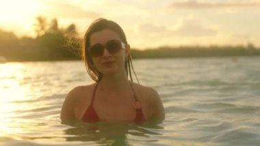 Sakin bir günbatımı sahnesinde bikinili bir kadın okyanusun tadını çıkarıyor, zarif bir şekilde suda duruyor ve nazik dalgalarda yüzüyor.