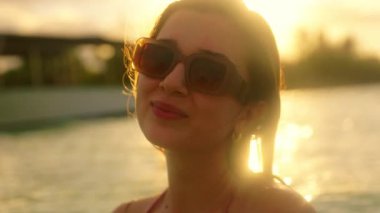Bu video, güneş gözlüklü neşeli bir kadını, huzur ve mutluluğu somutlaştıran, deniz kenarında gün batımının dingin güzelliğini gözler önüne seriyor.
