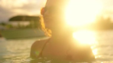 Büyüleyici bir günbatımı sahnesi, sığ suda yüzen ve güneş gözlükleriyle renkleri yansıtan bikinili bir kadın tasvir ediyor. Rüya gibi bir alacakaranlıkta özgürlüğünü somutlaştırırken dalgalar etrafında döner.