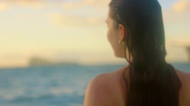 Huzurlu bir sahil sahnesinde bikinili bir kadın okyanusun tadını çıkarıyor. Suda duruyor, kameraya bakıyor ve gün batımında neşeli oluyor.