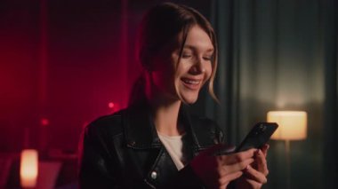 Genç bir kadın şık ve canlı bir atmosferde akıllı telefonunu kullanırken gülümsüyor, neşe ve bağlantı yayıyor..