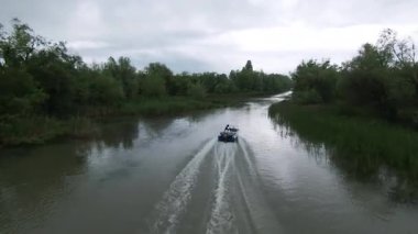 Vahşi kırsal nehir manzarasında yelkenli yelkenliyi takip ediyorum. Yapay zeka robotu ve erkek turistler teknede. Fütürist gezi konsepti. FPV İHA uçuşu.