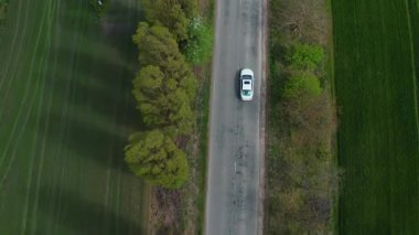 Kırsal alanda hızla ilerleyen beyaz modern arabanın havadan görünüşü. Kırsal görüşler ve yeşil doğa gelip geçiyor. Yolculuk, seyahat konsepti.