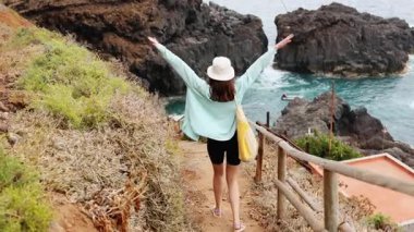 Deniz kenarındaki güzel tepelik uçurumlarda aşağı inerken kollarını uzatan mutlu özgür beyaz kadın. Karanlık kayalıkları olan harika bir kumsal..