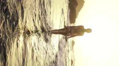 Gün batımında sakin turuncu deniz manzarasına doğru yürüyen güzel seksi kadın siluetinin dikey görüntüsü. Sinema kumsalındaki çekici kız..