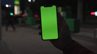 NEW YORK - 19 Nisan 2019: Elinde tuttuğu adam, gece sehrinin yeşil arka planında dikey yeşil ekranlı akıllı telefonu kullanıyor.