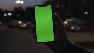 NEW YORK - 19 Nisan 2019: Elinde akıllı telefonu olan ve gece şehir renklerinde yeşil ekranlı akıllı telefonu kullanan bir adam dijital cihaz, mobil dokunmatik, mobil, yavaş çekimde araç iletişimi cihazını teslim etti