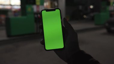 NEW YORK - 19 Nisan 2019: Elle tutulan eller, gece şehrinin yeşil arka planında dikey yeşil ekranlı akıllı telefonu kullanıyor.