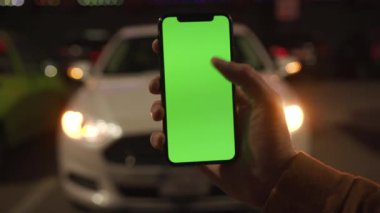 NEW YORK - 19 Nisan 2019: Genç adam elinde dikey yeşil ekranlı akıllı telefon kullanıyor ve gece renkli arka plan beyaz araba iletişimi cihazı dijital cihaz mobil dokunmatik mobil yavaş çekim