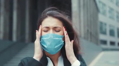 Sağlıksız, üzgün, beyaz, tıbbi maske takan ve baş ağrısı çeken genç bir kadının portresi sokakta şakaklarına dokunuyor. Karantina.