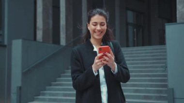 Binanın dışında akıllı telefon uygulaması kullanarak yürüyen genç bir bayanın sosyal medyada neşeli ve rahat bir şekilde mesajlaşması..