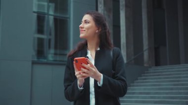 Genç neşeli ofis kızı Kafkasyalı güzel kadın dışarıda iş alanında yürürken akıllı telefon ağı sosyal medya kullanıyor. Kaygısız. Kentsel yaşam tarzı.