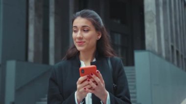 Neşeli, genç, yetişkin, beyaz kadın ofis müdürü modern binanın yanında yürüyor mutlu bir şekilde haberlere bakıyor, akıllı telefon sosyal medyası açık havada..