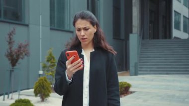 Kafası karışmış beyaz bir kadın akıllı telefondan haber zaman çizelgesi okuyor. Şaşırmış bir yüz ifadesiyle ofis binasının önünde durmakta tereddüt ediyor..