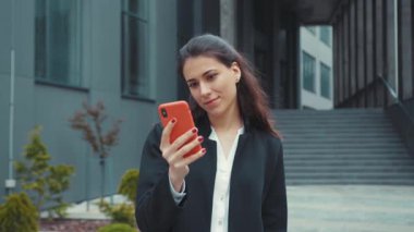 Neşeli, genç, çekici, beyaz kadın modern ofis binasının yanında duruyor. Akıllı telefondan mesaj yazıyor, gülümsüyor, sosyal medya zaman çizelgesini tarıyor..