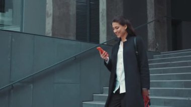 Akıllı telefon kullanarak ofisten ayrılan Kafkasyalı güzel genç bir iş kadını dışarıda mutlu zaman çizelgesi sosyal medyası okuyor. Neşeli insanlar. Kentsel yaşam tarzı.