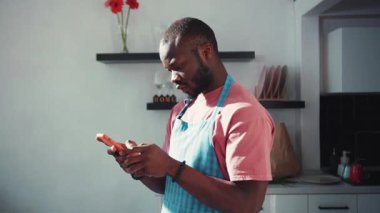 Afro-Amerikalı çekici bir adam yemek pişirirken internette geziniyor. Akıllı telefondan mesaj aldığında gülümseyen neşeli siyahi adam. İç mekanlar. Ev hayatı. Teknoloji.