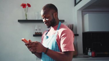 Afro-Amerikalı önlüklü bir adam mutfaktaki akıllı telefon sosyal medya zaman çizelgesinde geziniyor. Sıradan bir yaşam tarzı. - Evde kalıyorum. İnsanlar ve teknoloji.