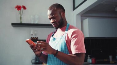 Önlük giyen bir adam akıllı telefonuyla meşgul olurken parlak, davetkar bir mutfakta yemek pişiriyor.