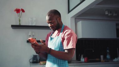 Önlük takan bir adam modern bir mutfakta aynı anda lezzetli yemekler pişirirken telefonunu kontrol ediyor.